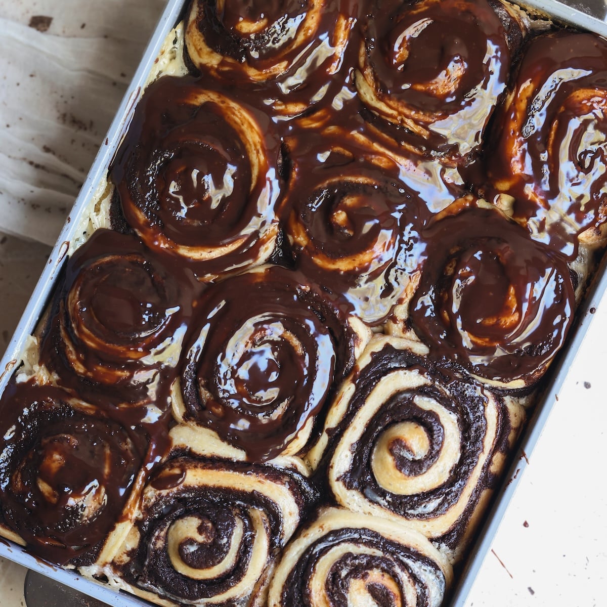 A pan of brownie cinnamon rolls, some topped with glossy chocolate glaze and others plain, rests on a light-colored surface.