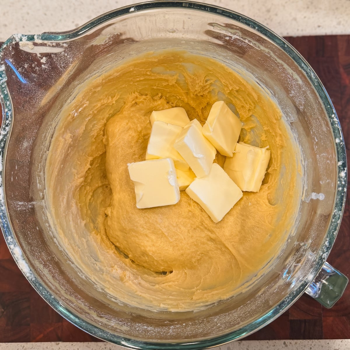 A glass mixing bowl with thick dough and several chunks of butter on top, sitting on a wooden surface.