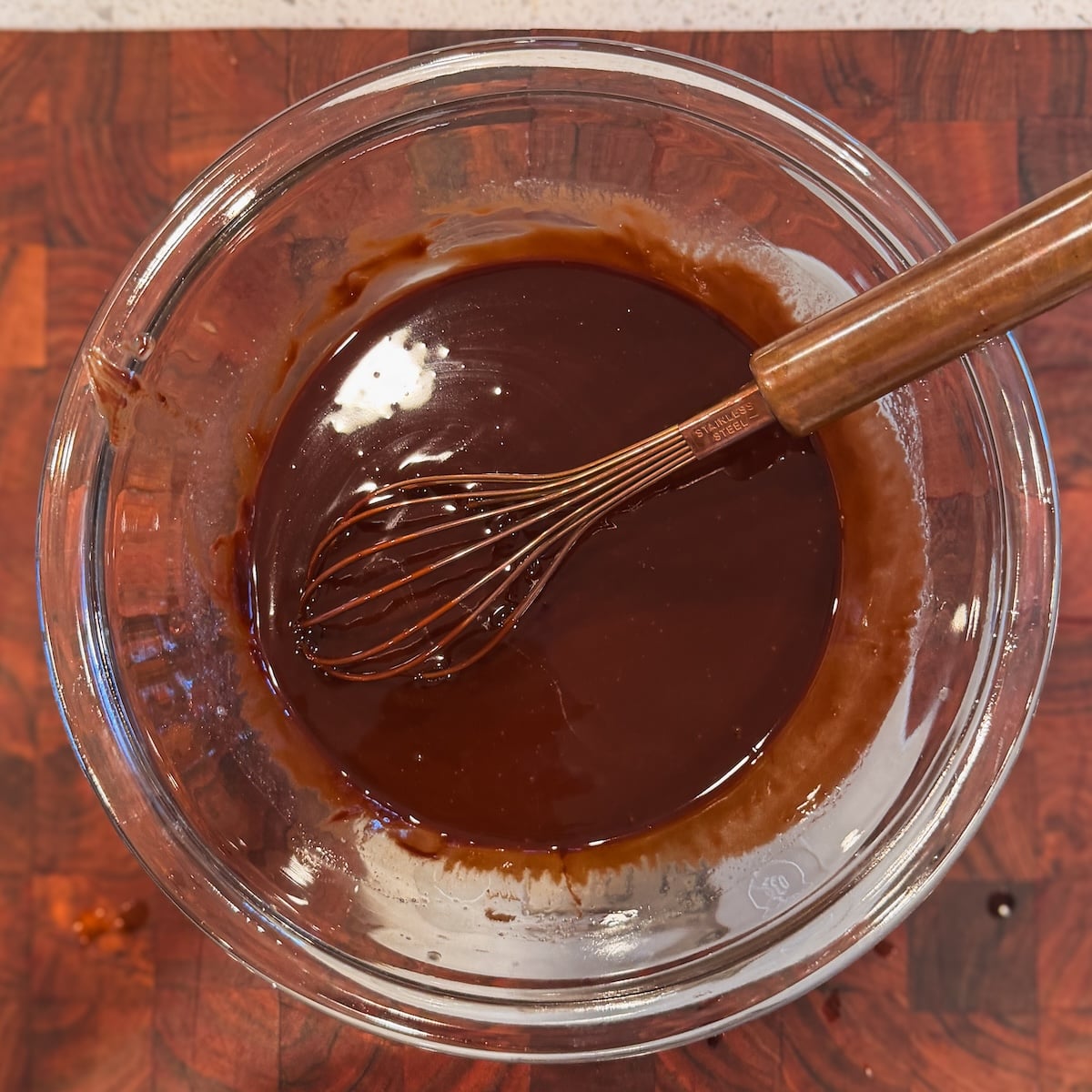 A glass bowl filled with melted chocolate and butter being mixed with a whisk, placed on a wooden surface.