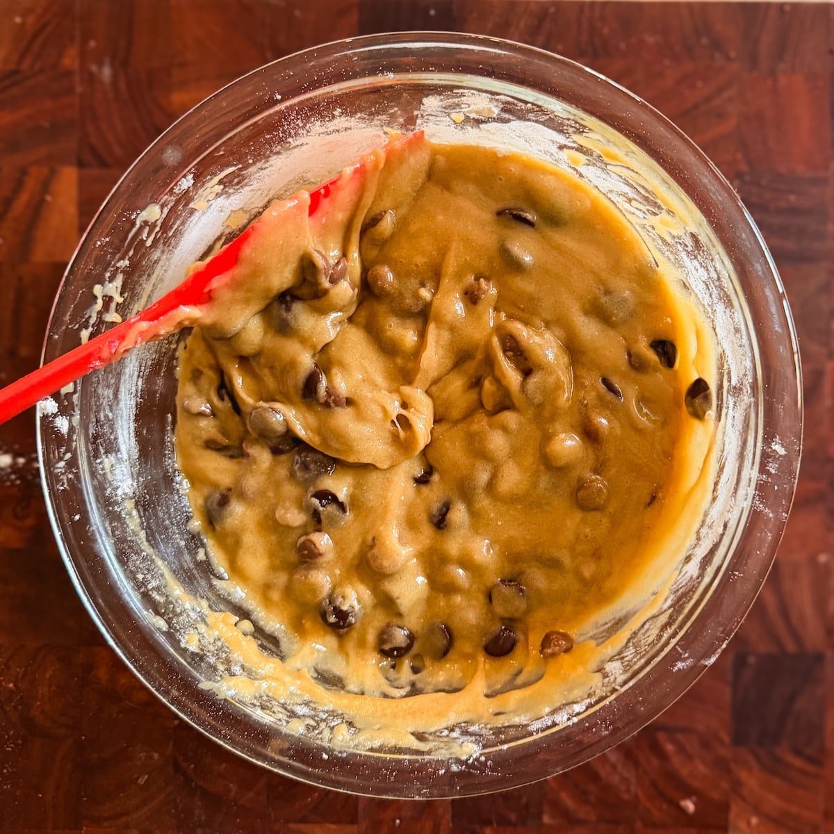 A glass bowl filled with chocolate chip cookie dough and a red spatula resting on the side, sitting on a wooden surface.