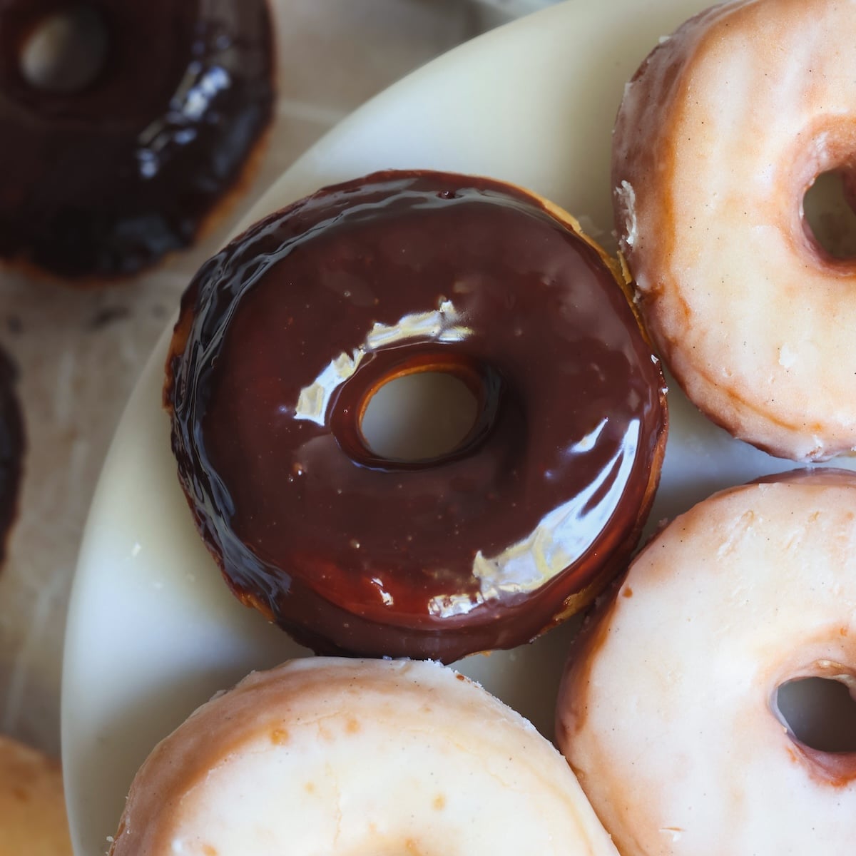 A close-up of glazed donuts on a white plate, with one donut covered in chocolate glaze and the others in a light sugar glaze.