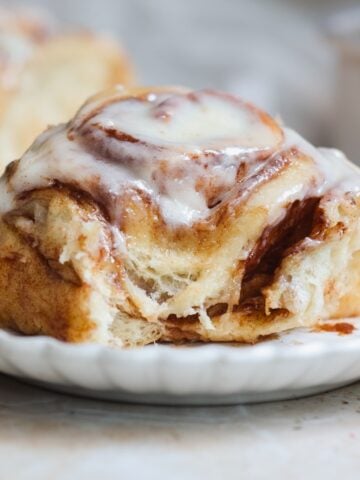A close-up of a cinnamon roll topped with creamy white icing sits on a white plate, with another cinnamon roll blurred in the background. It looks soft and gooey inside