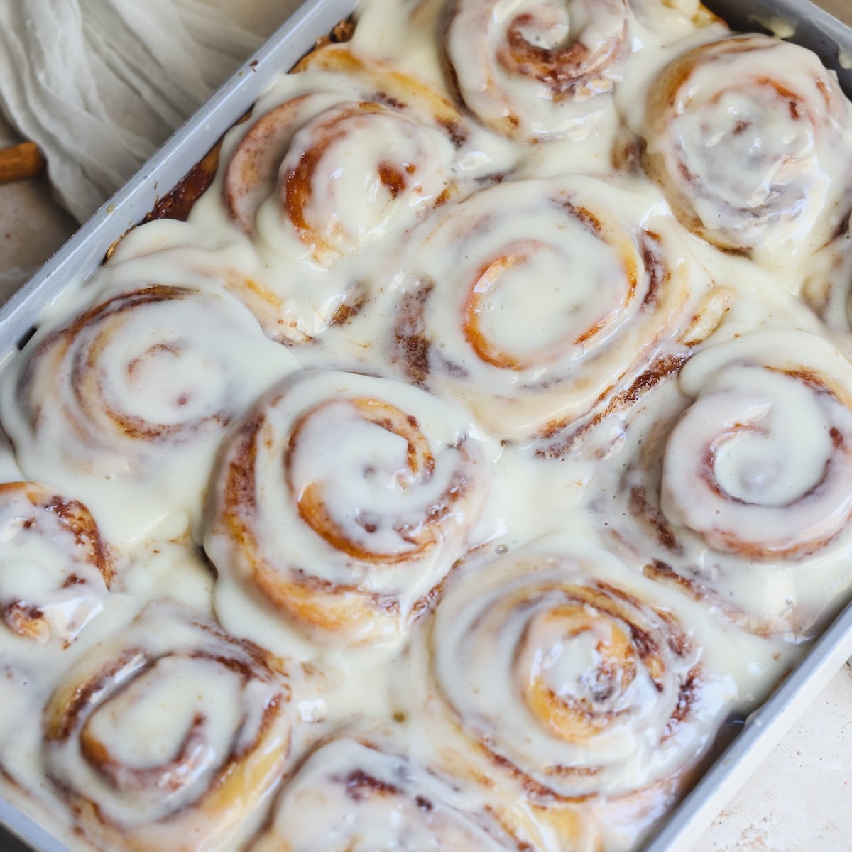 A baking tray filled with freshly baked cinnamon rolls, generously topped with creamy white icing that is slightly melted.