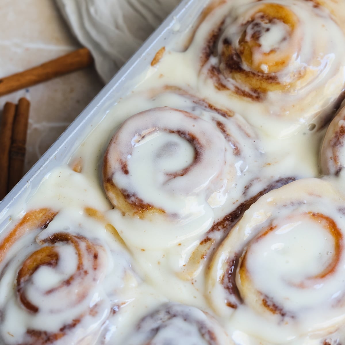 Close-up of freshly baked cinnamon rolls in a baking dish, drizzled generously with creamy white icing. Two cinnamon sticks and a napkin are visible in the background.