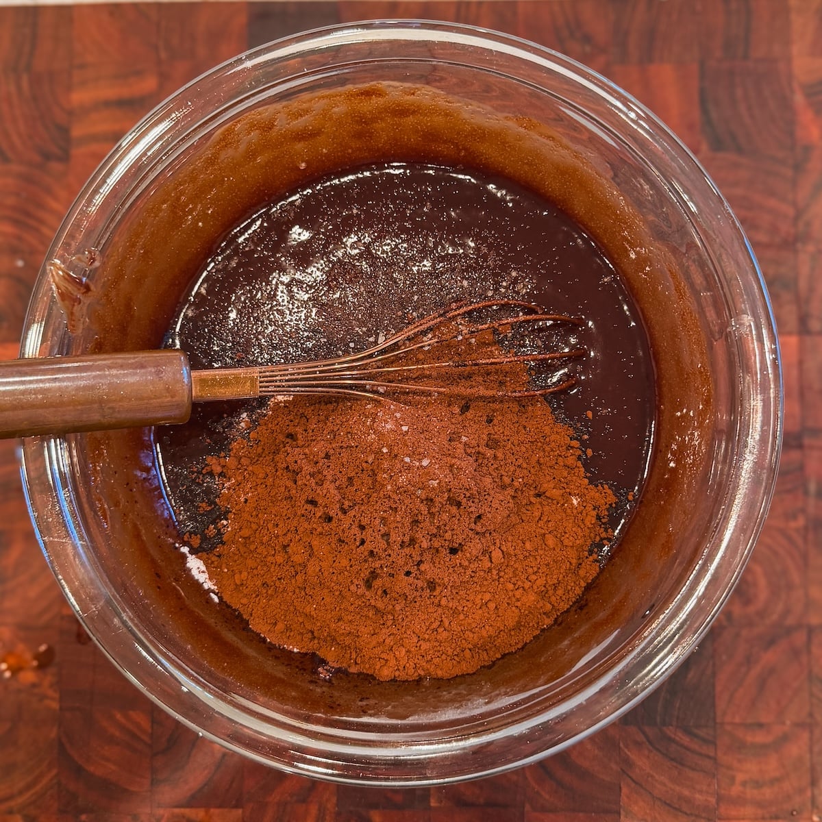 A glass bowl filled with chocolate mixture and cocoa powder being whisked, sitting on a wooden surface.