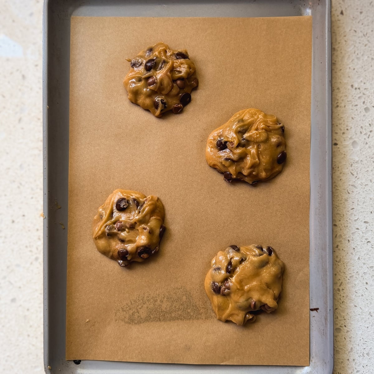 Four unbaked chocolate chip cookie dough mounds are spaced apart on a parchment-lined metal baking sheet, resting on a light-colored countertop.