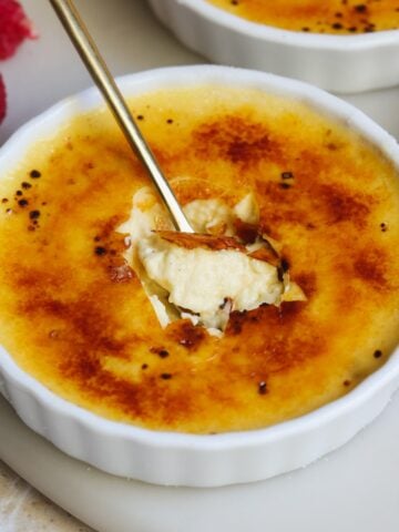 A spoon breaks through the caramelized sugar crust of a crème brûlée in a white ramekin, revealing creamy custard inside. Fresh raspberries are visible in the background.