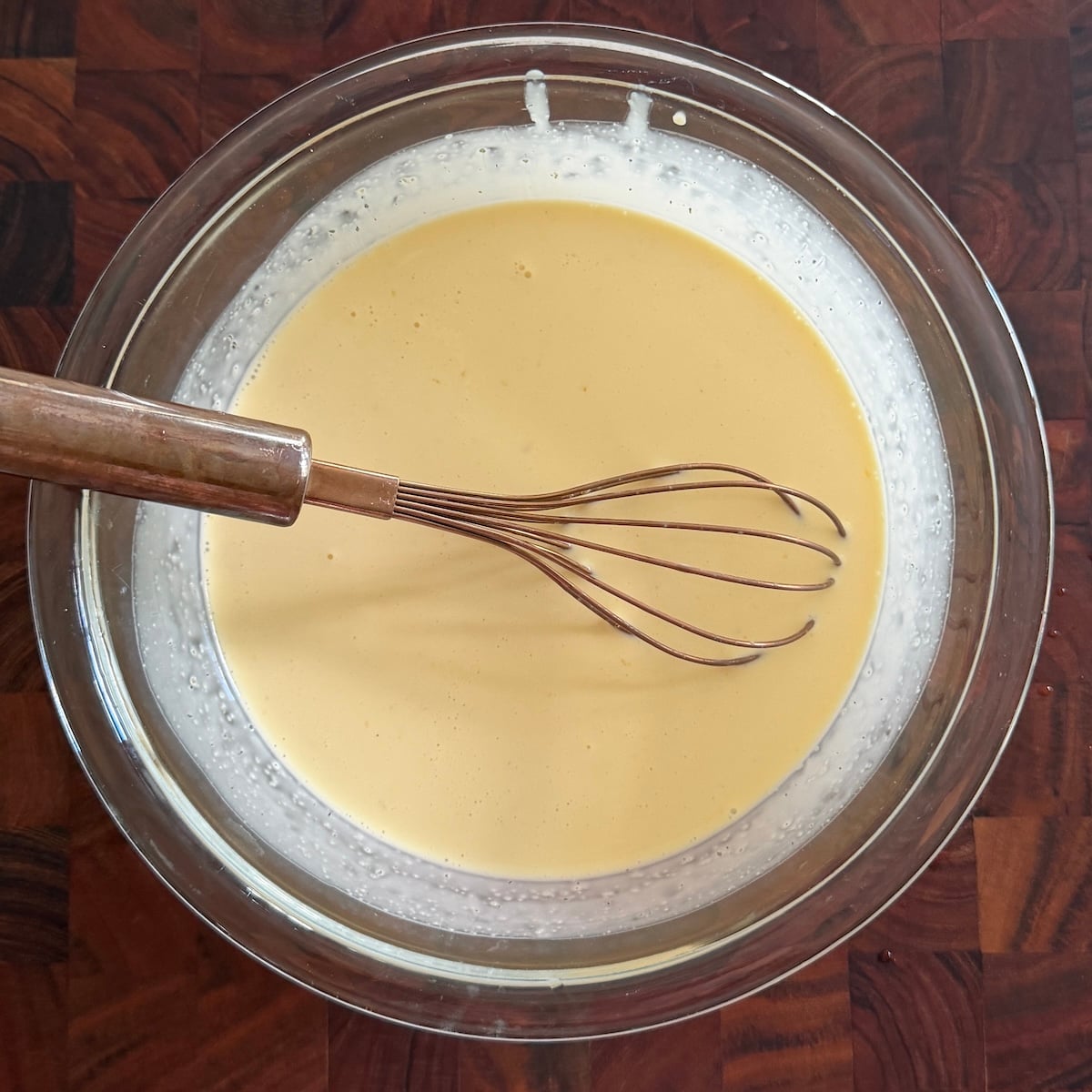A metal whisk rests in a glass bowl filled with smooth, pale yellow custard on a dark wooden surface.