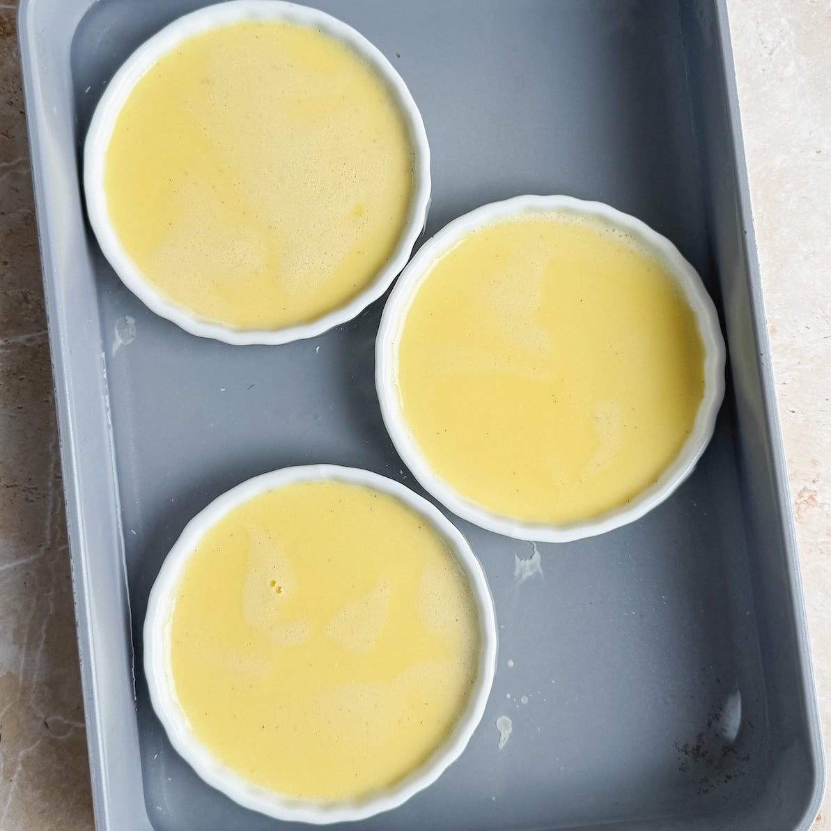 Three round white ramekins filled with a yellow custard creme brulee mixture are placed in a gray baking tray on a light-colored surface.