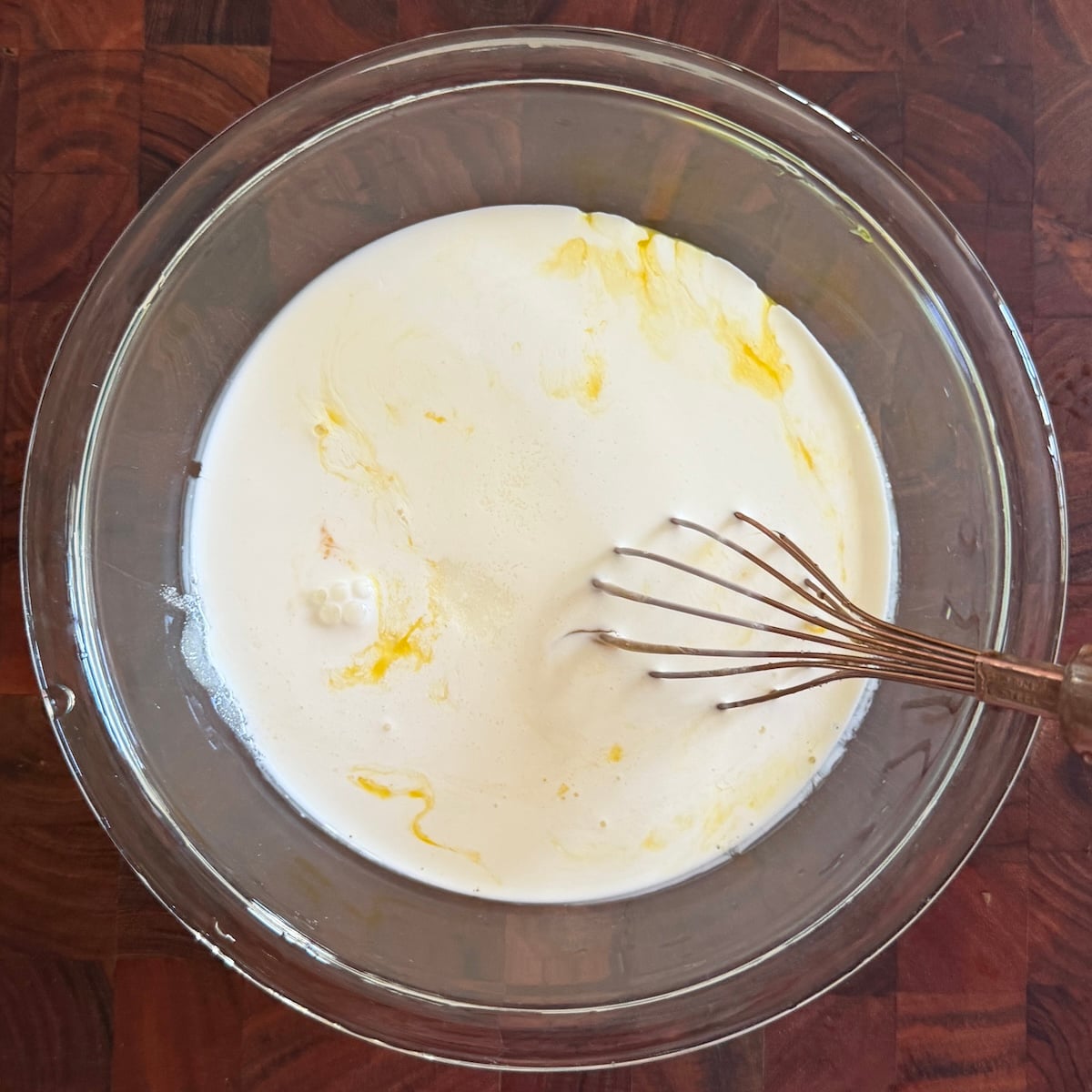 A glass bowl with cream, egg yolks, and a whisk on a wooden surface. Some yellow egg yolk is swirling into the cream, and a small frothy spot appears near the edge.
