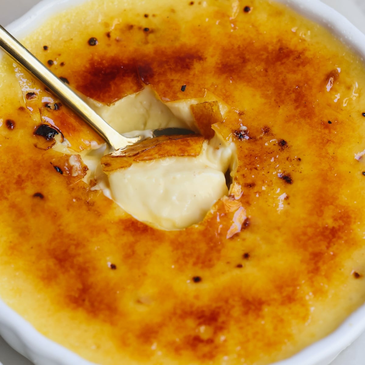 A close-up of crème brûlée in a white dish, with a golden caramelized sugar crust cracked open by a spoon, revealing creamy custard underneath.