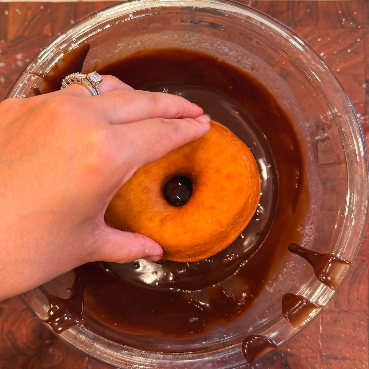 A hand with a diamond ring dips a plain donut into a bowl of chocolate glaze, with some chocolate dripping down the side of the bowl.
