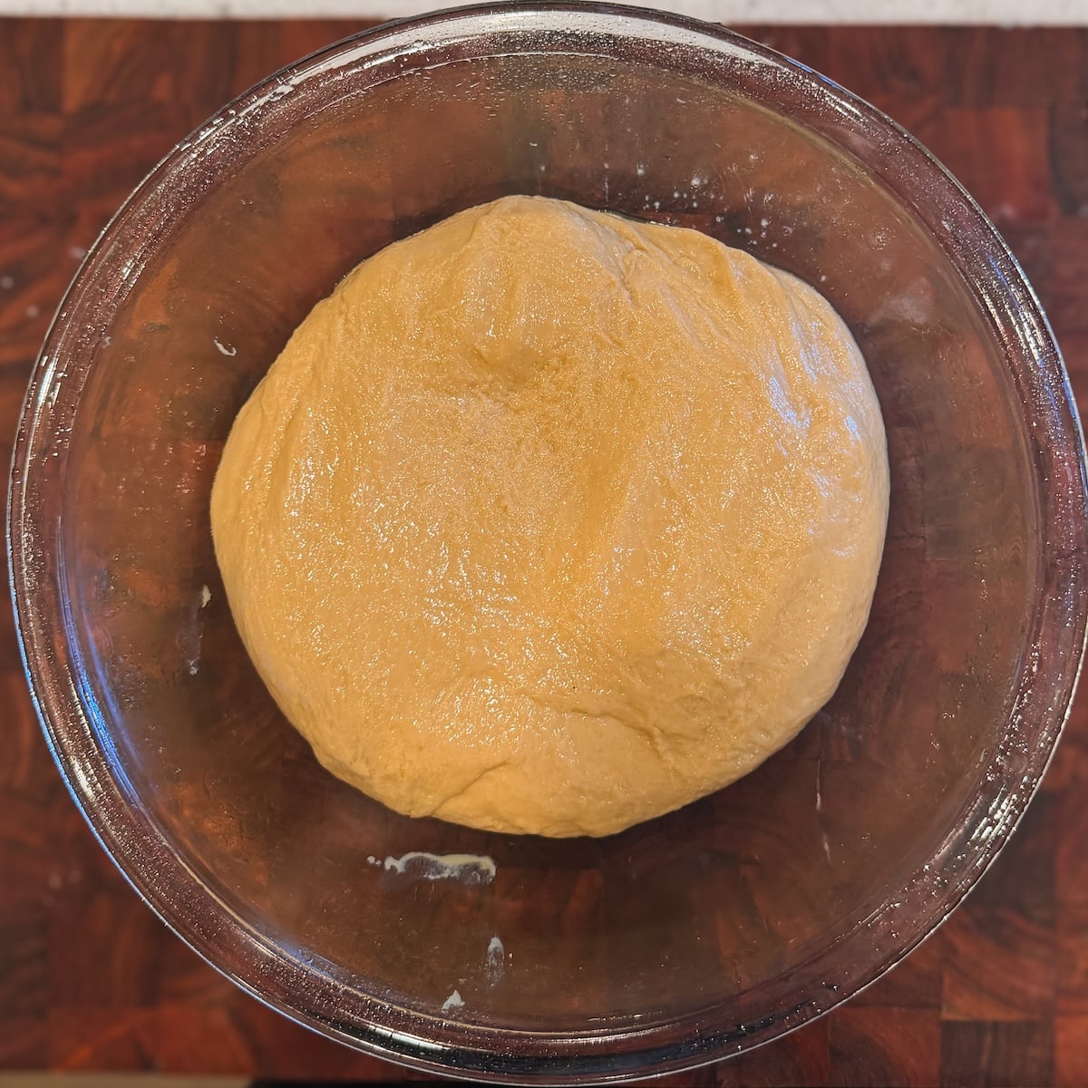A ball of dough rests in a clear glass bowl on a dark wooden surface, ready to rise.