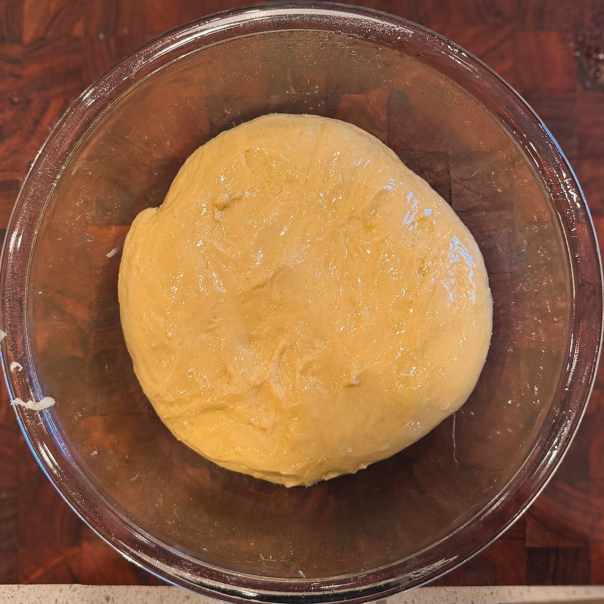 A round ball of yellow dough sits in a clear glass bowl on a dark wooden surface, ready to rise or be shaped.