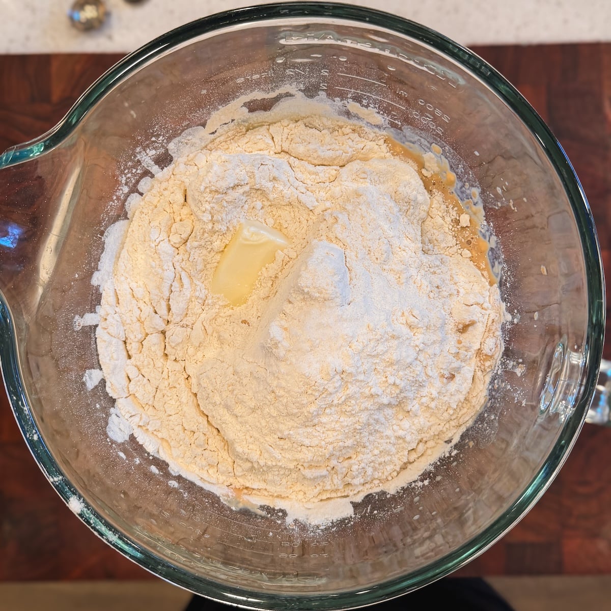 A glass bowl contains flour, a pat of butter, and other dry baking ingredients, viewed from above on a wooden surface.