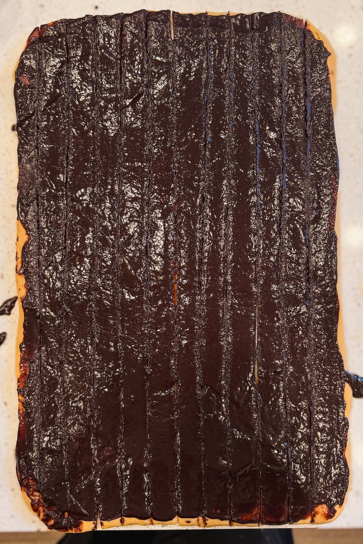 A rectangular pastry covered in a thick, glossy layer of brownie batter, with visible vertical knife marks dividing it into narrow strips. The pastry sits on a light-colored surface.