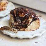 A chocolate-swirl cinnamon roll topped with melted chocolate sits on a white scalloped plate. A gold fork with chocolate smudges rests beside the pastry.