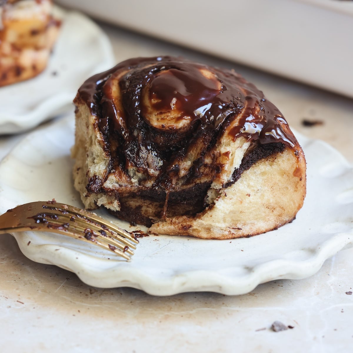A chocolate-swirl cinnamon roll topped with melted chocolate sits on a white scalloped plate. A gold fork with chocolate smudges rests beside the pastry.