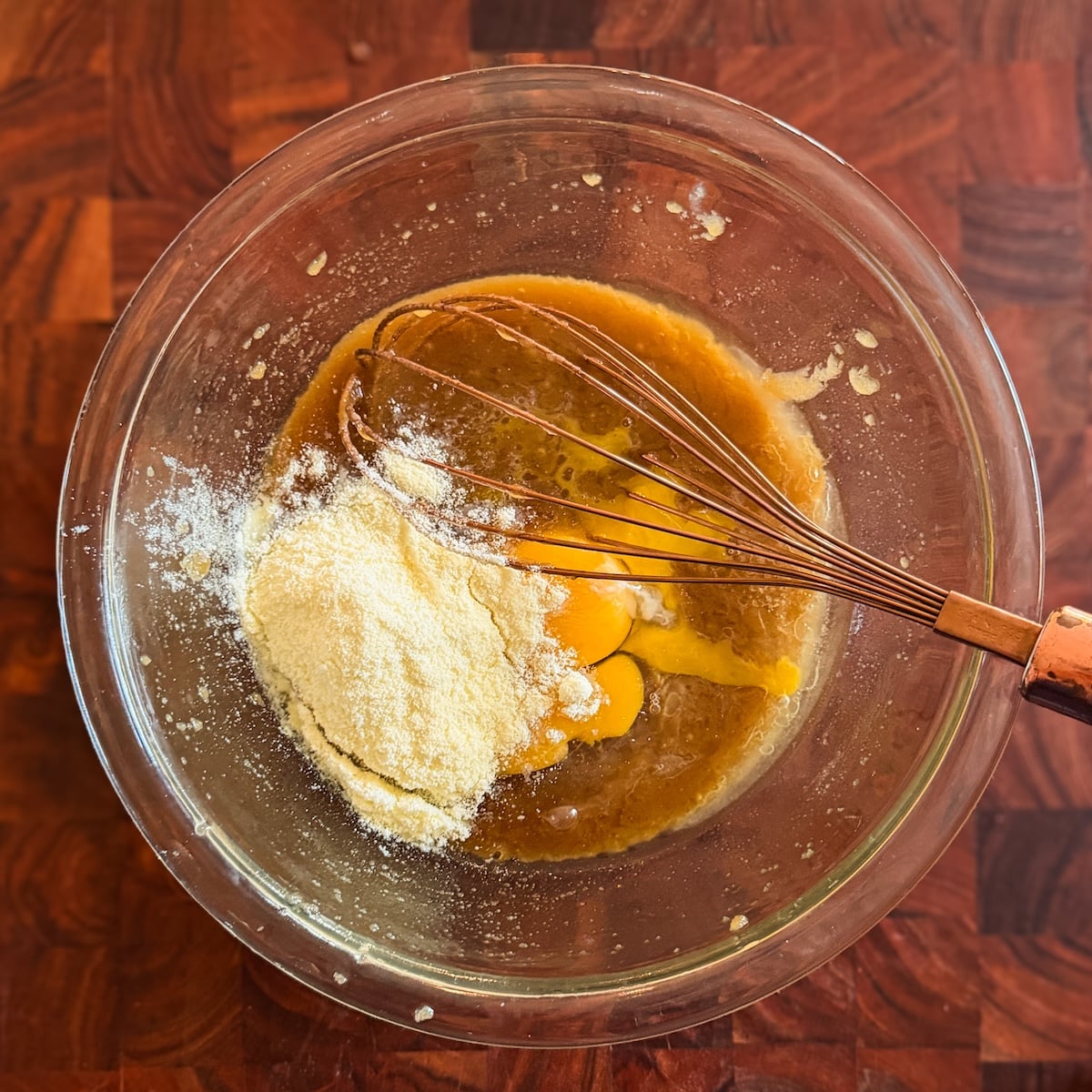 A glass bowl on a wooden surface holds eggs, milk powder, and wet ingredients being mixed with a metal whisk.