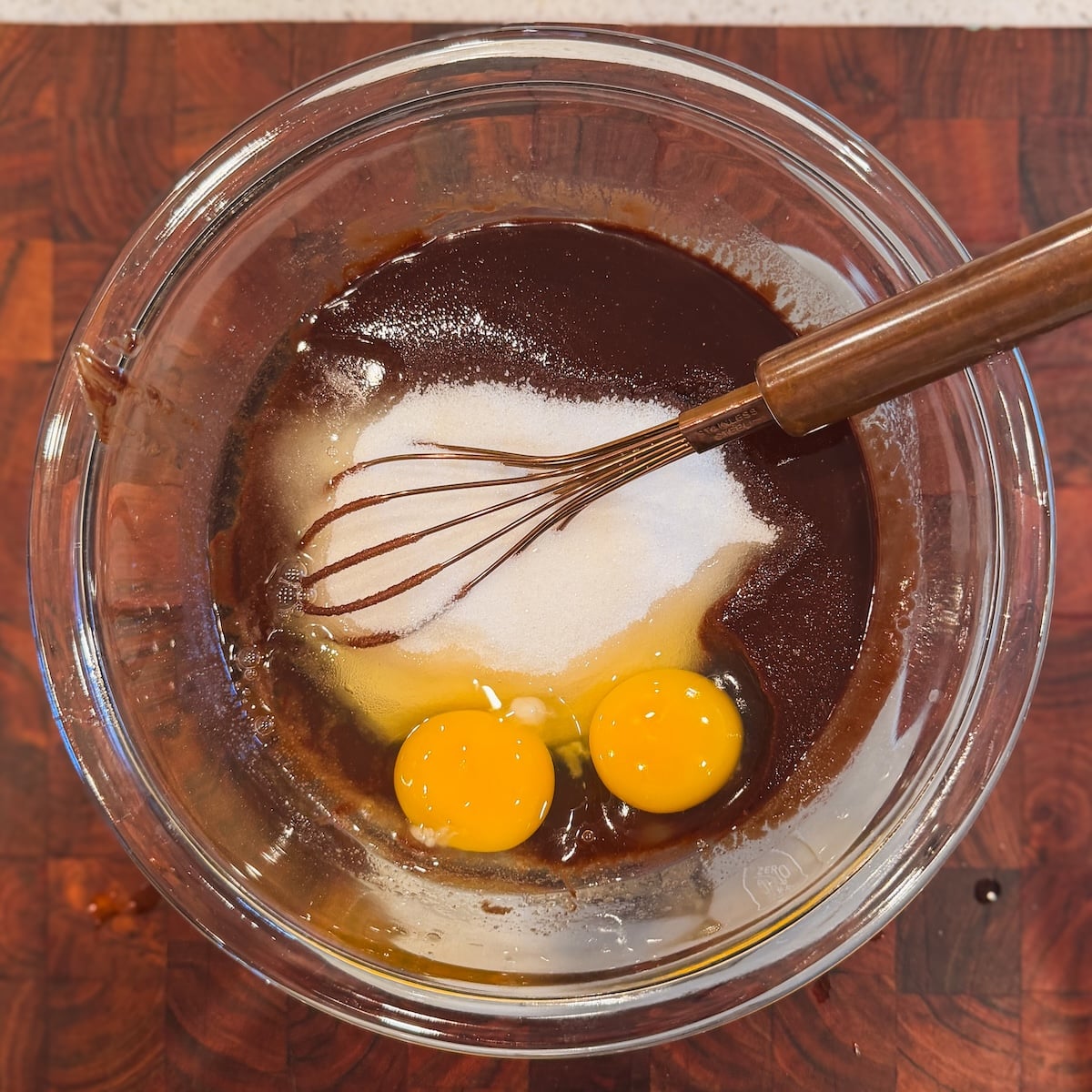 A glass bowl with melted chocolate, two eggs, and sugar, being mixed with a metal whisk on a wooden surface.