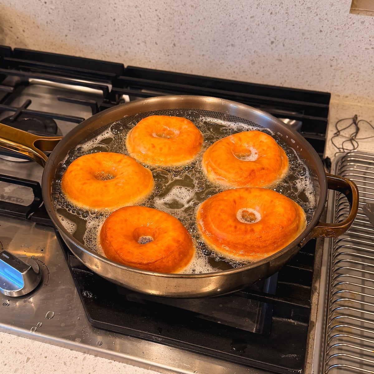 Five doughnuts are frying in oil in a large pan on a stovetop, turning golden brown. A cooling rack sits nearby on the countertop.