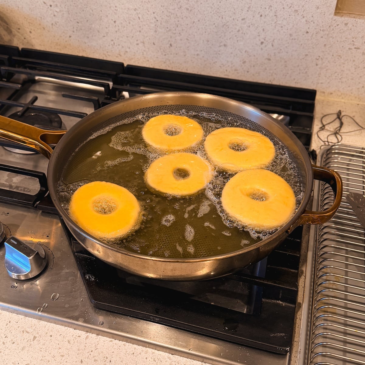 Six donut-shaped pieces of dough are frying in hot oil in a large pan on a stovetop, with bubbles forming around them. A cooling rack is placed nearby on the counter.