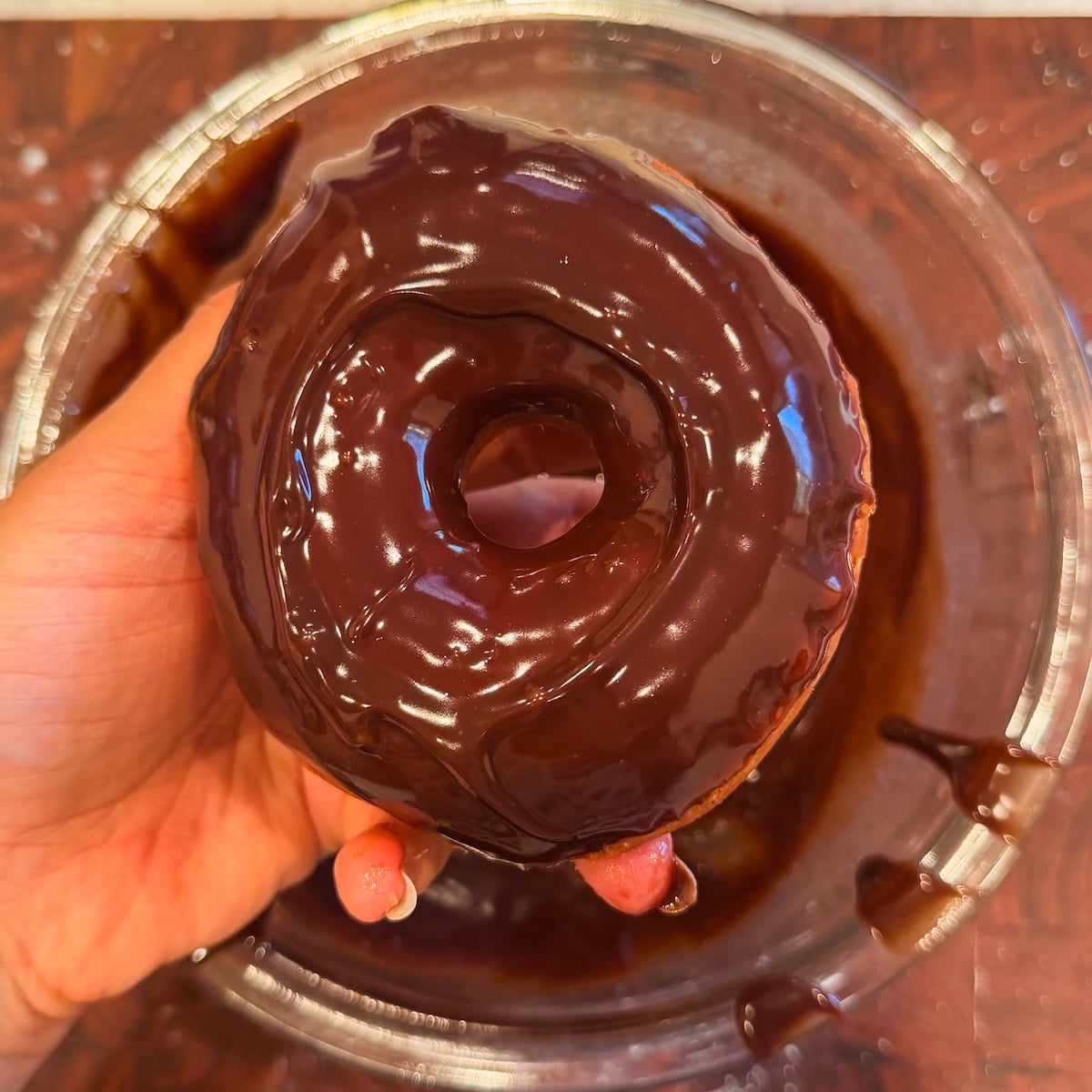 A hand holding a donut freshly dipped in glossy chocolate glaze over a glass bowl with chocolate drips.