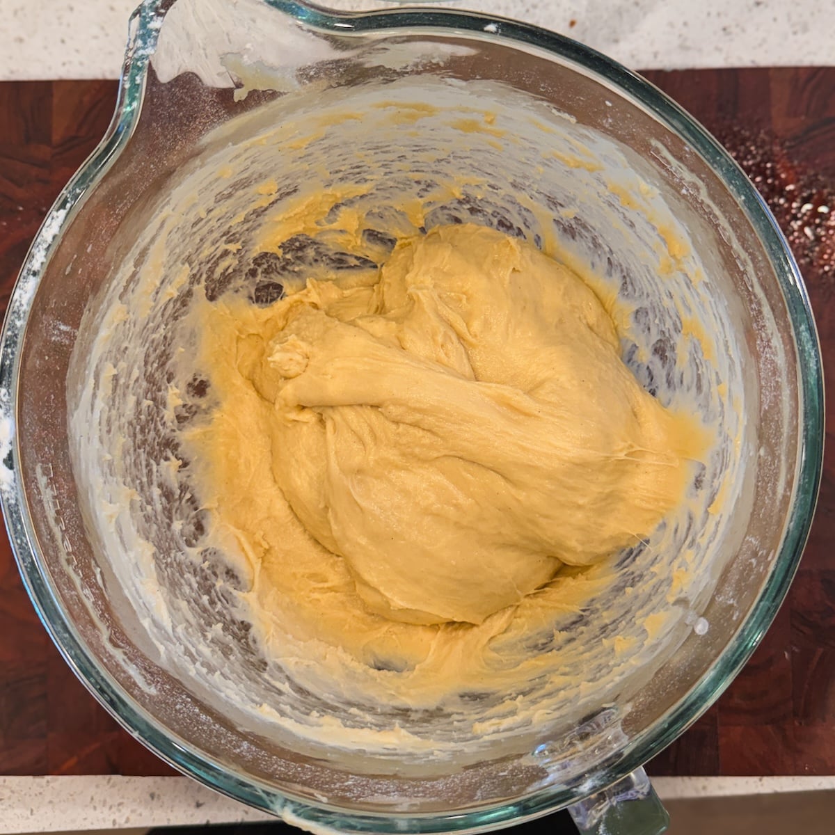 A glass mixing bowl filled with thick, yellow dough sits on a dark wooden cutting board, with some flour scattered in the background.