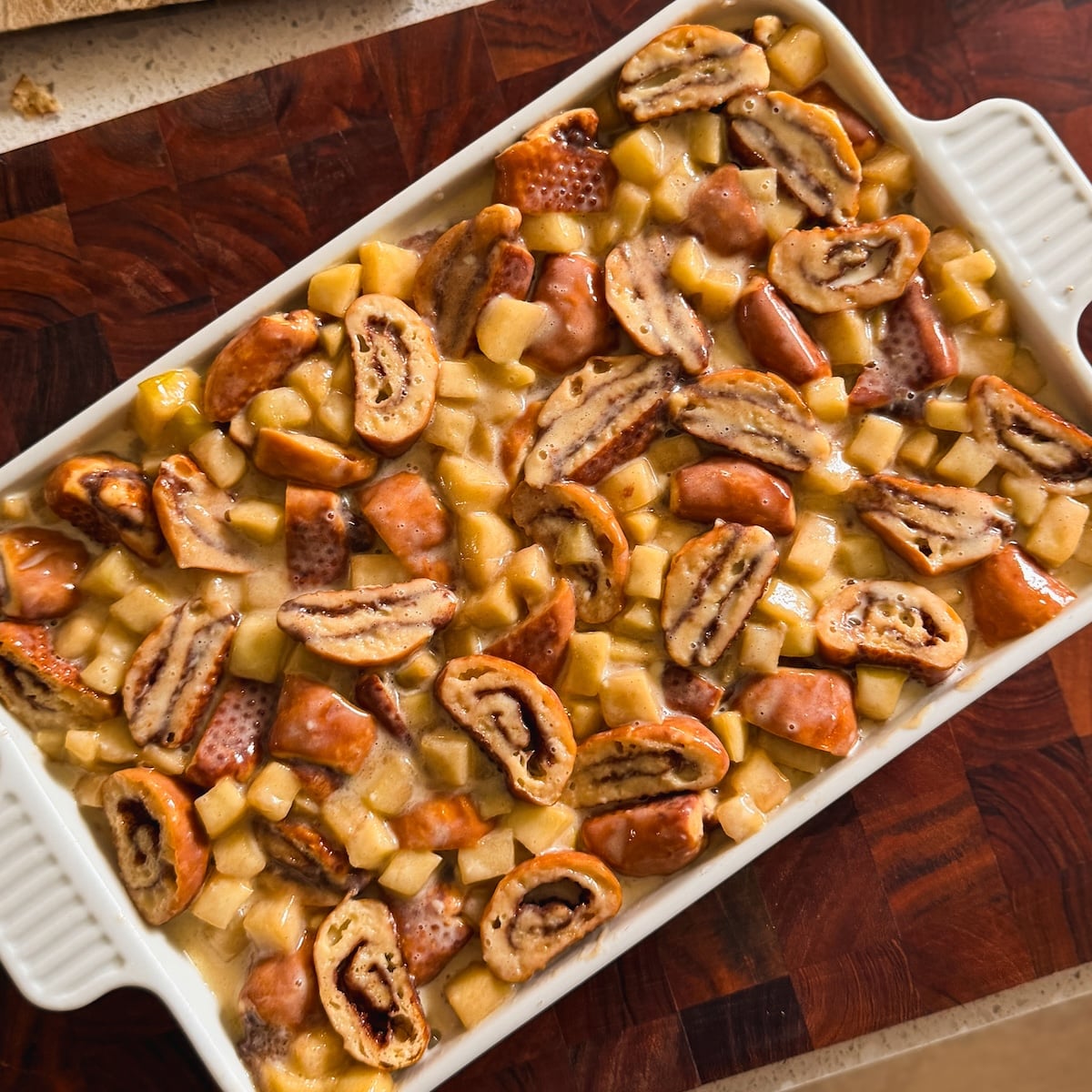 A white baking dish holds a dessert with diced apples and sliced cinnamon roll bites, soaked in a custard ready for baking