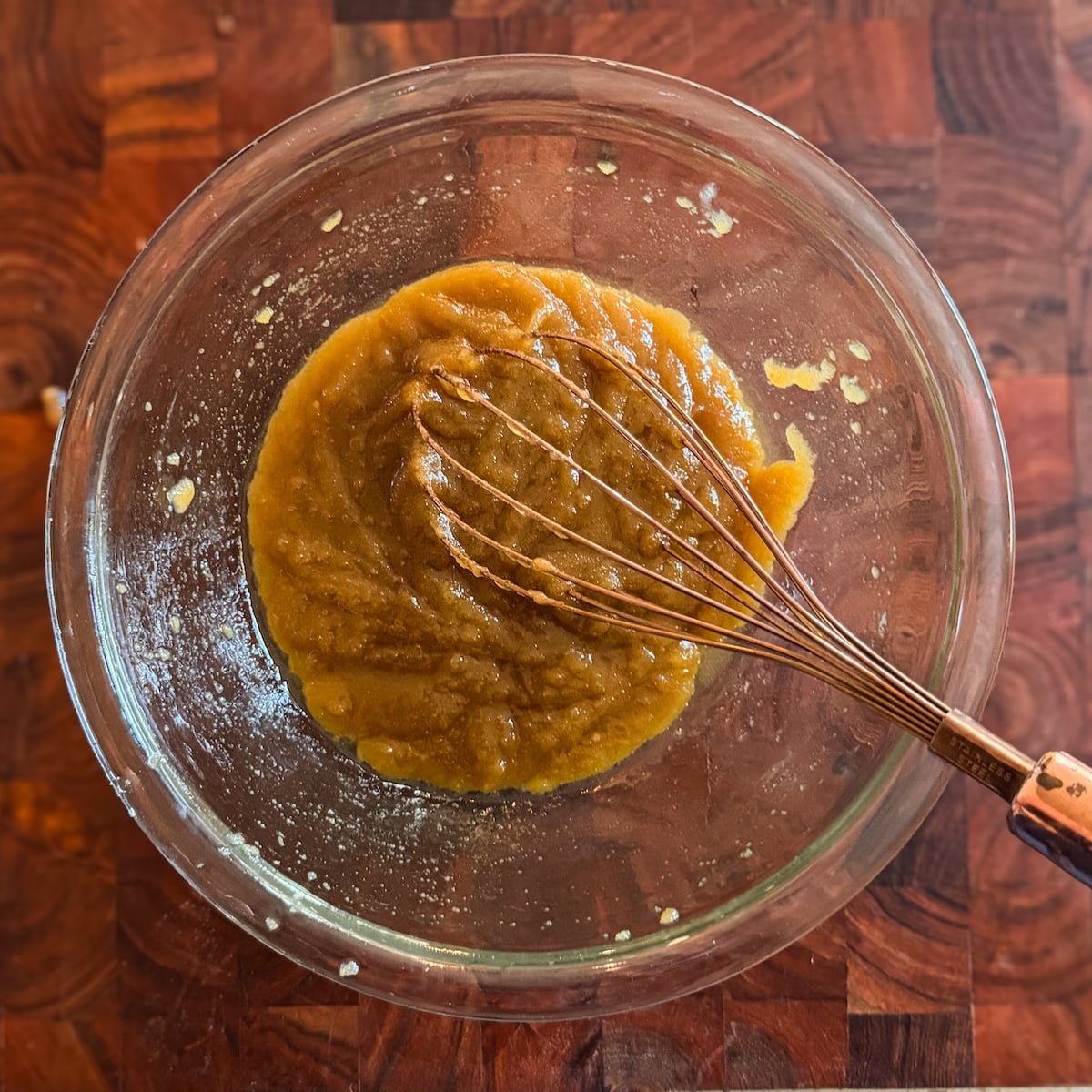 A glass bowl with thick brown mixture of sugar and butter being whisked, placed on a wooden surface.