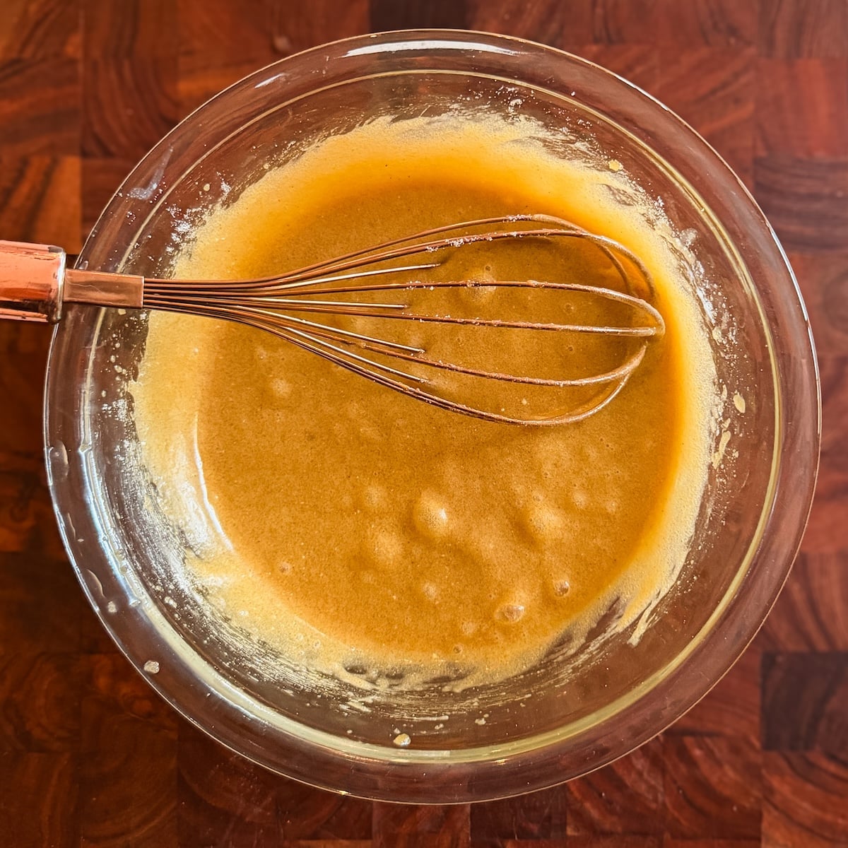 A glass bowl filled with light brown mixture of butter, sugar and egg sits on a wooden surface. A metal whisk with a copper handle rests inside the bowl, partially coated with the mixture.
