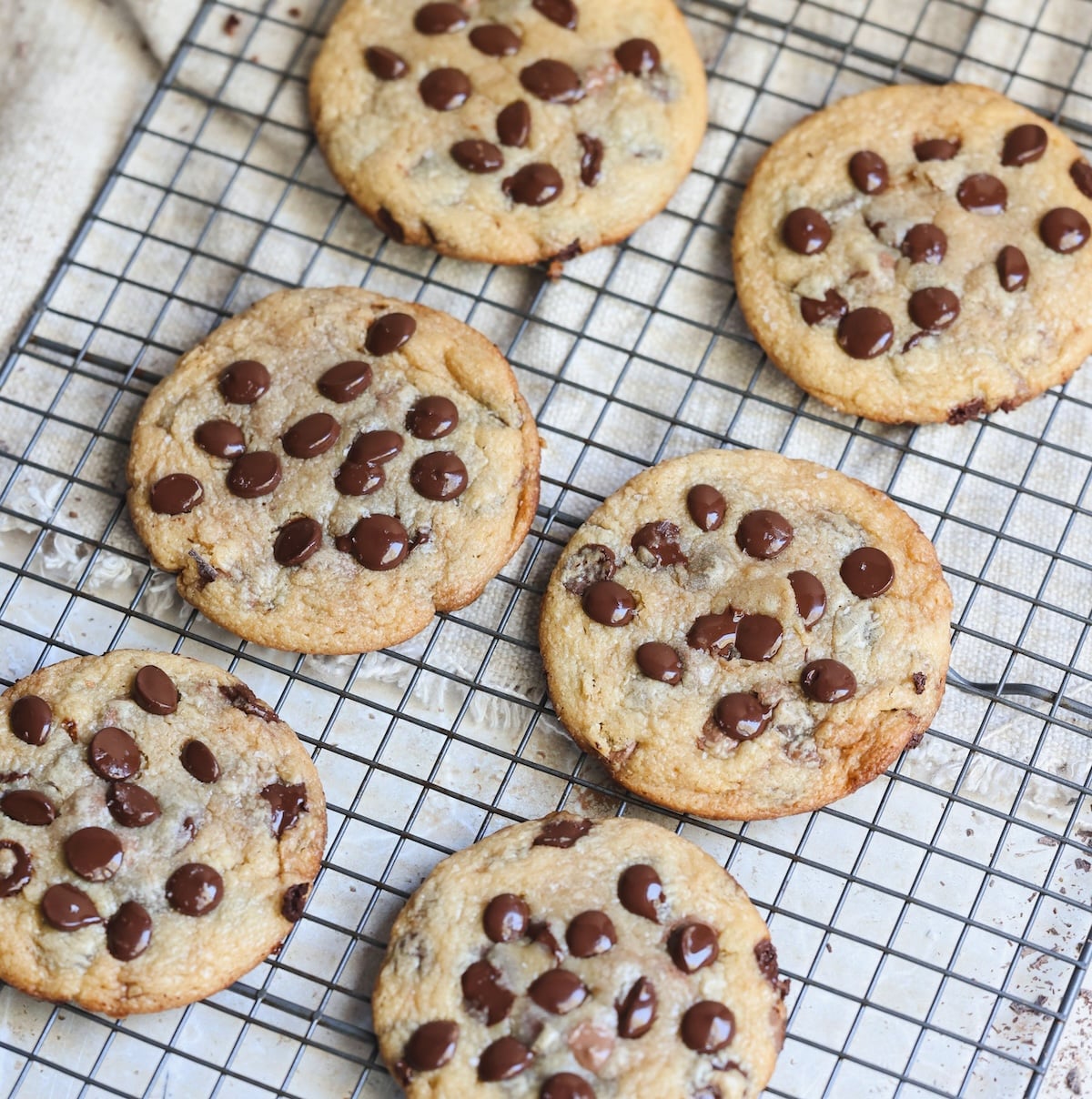 Six chocolate chip cookies cool on a wire rack. The cookies are golden brown with visible chocolate chips and have a slightly crisp edge. The rack sits on a lightly textured, light-colored surface.