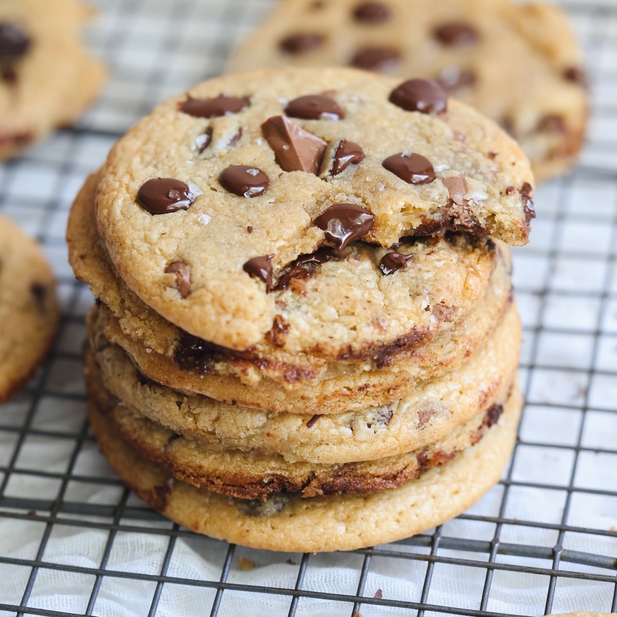 A stack of chocolate chip cookies on a cooling rack, with the top cookie missing a bite. Melted chocolate chips are visible on the cookies.