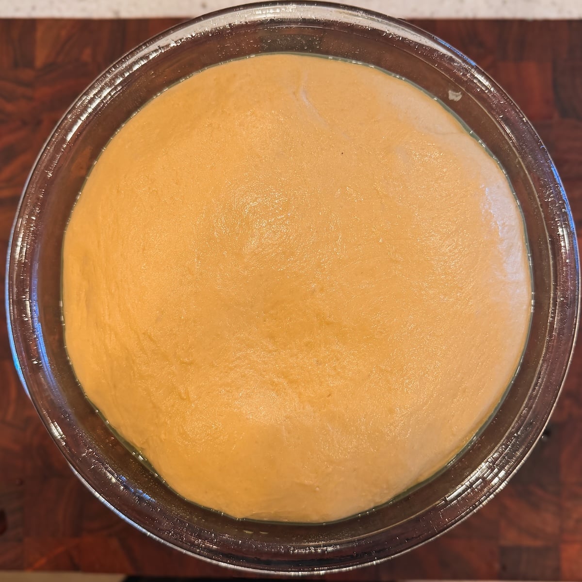 A glass bowl filled with risen dough sits on a wooden surface, showing a smooth, slightly domed top, ready for the next step in baking.