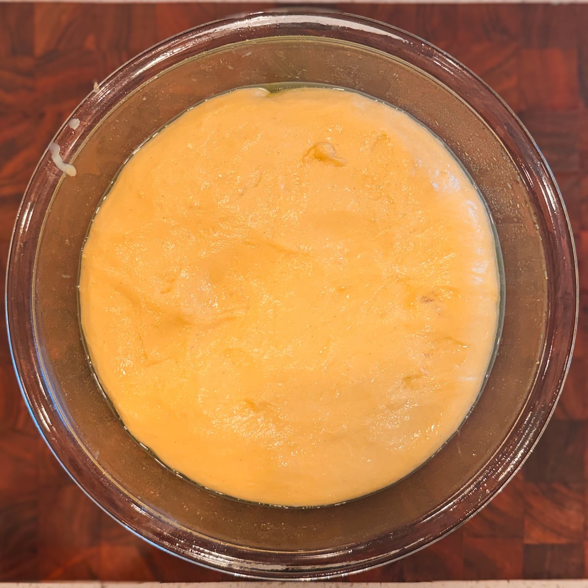 A glass bowl filled with risen dough sits on a wooden surface. The dough is smooth and slightly glossy, filling most of the bowl.