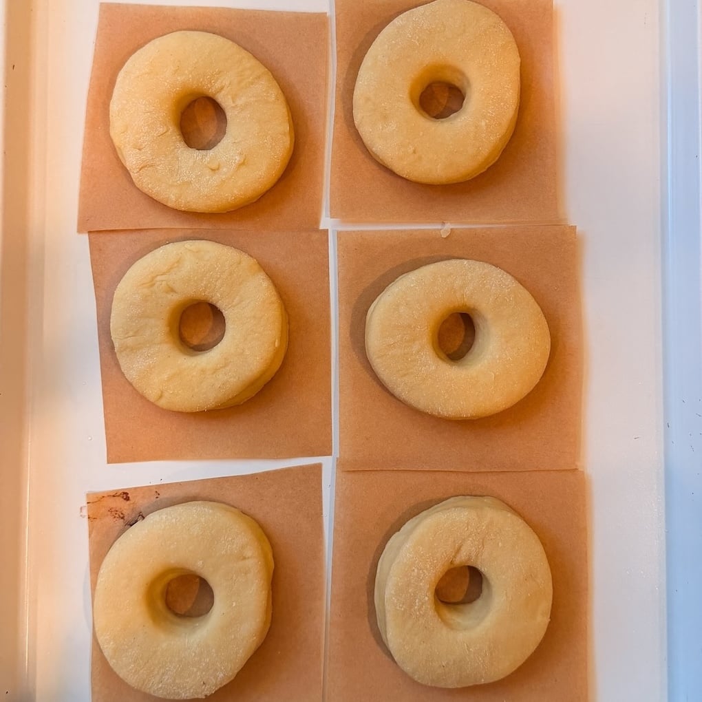 Six unbaked doughnuts are placed on individual squares of parchment paper, arranged in two columns and three rows on a white tray. They look puffy