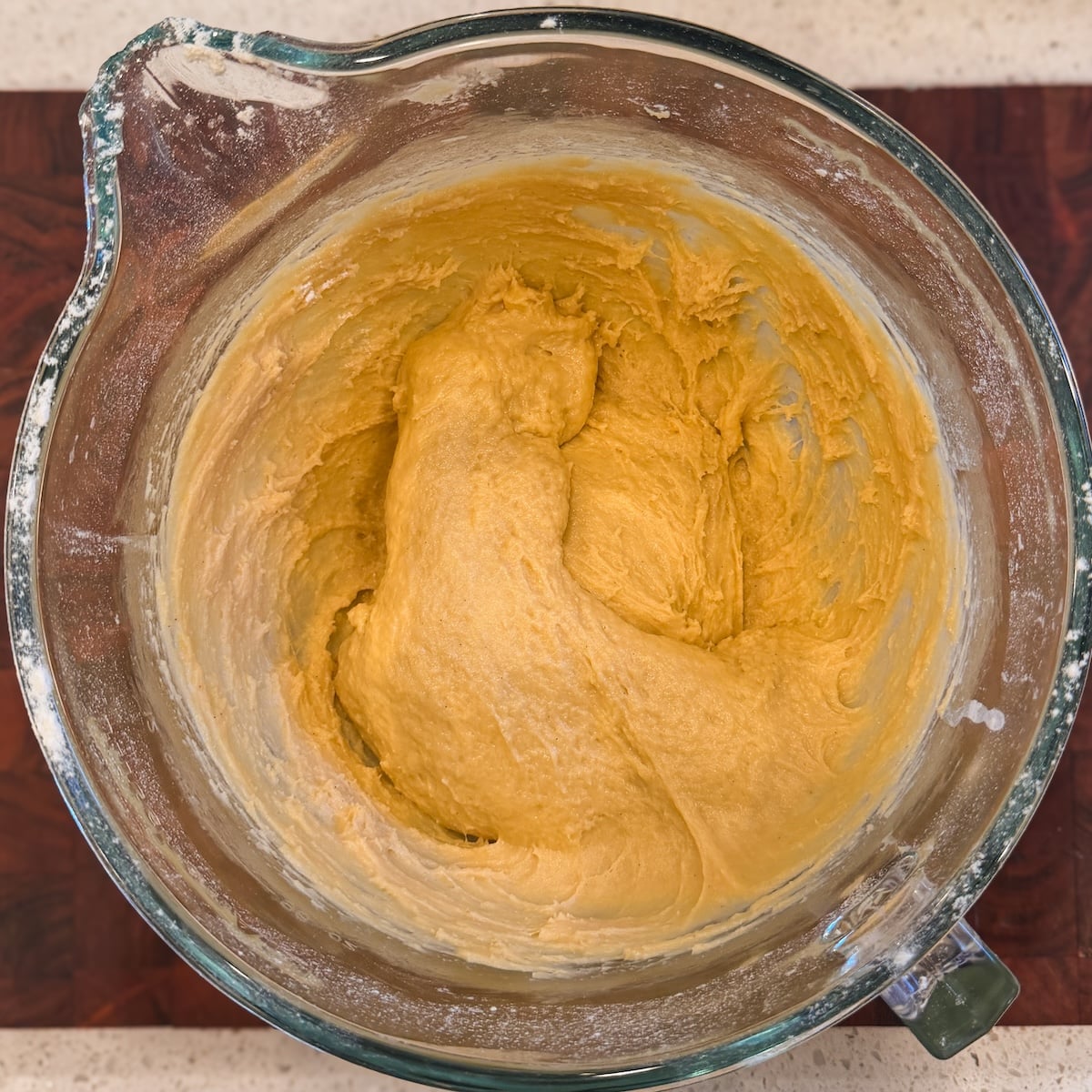A glass mixing bowl filled with smooth, golden-dough sits on a dark wooden surface. The dough appears freshly mixed, with some streaks left on the bowls sides.