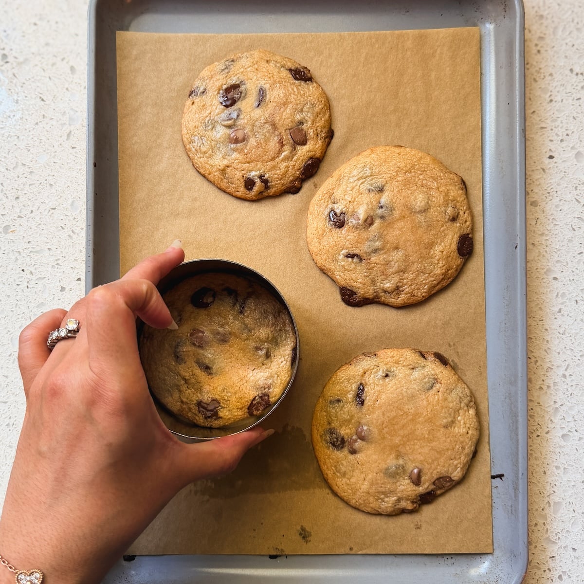 A hand holding a round metal cutter shapes a chocolate chip cookie on a parchment-lined baking tray with three other cookies.