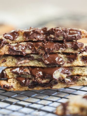 A stack of gooey chewy chocolate chip cookies, broken in half to show melted chocolate, sits on a wire cooling rack.