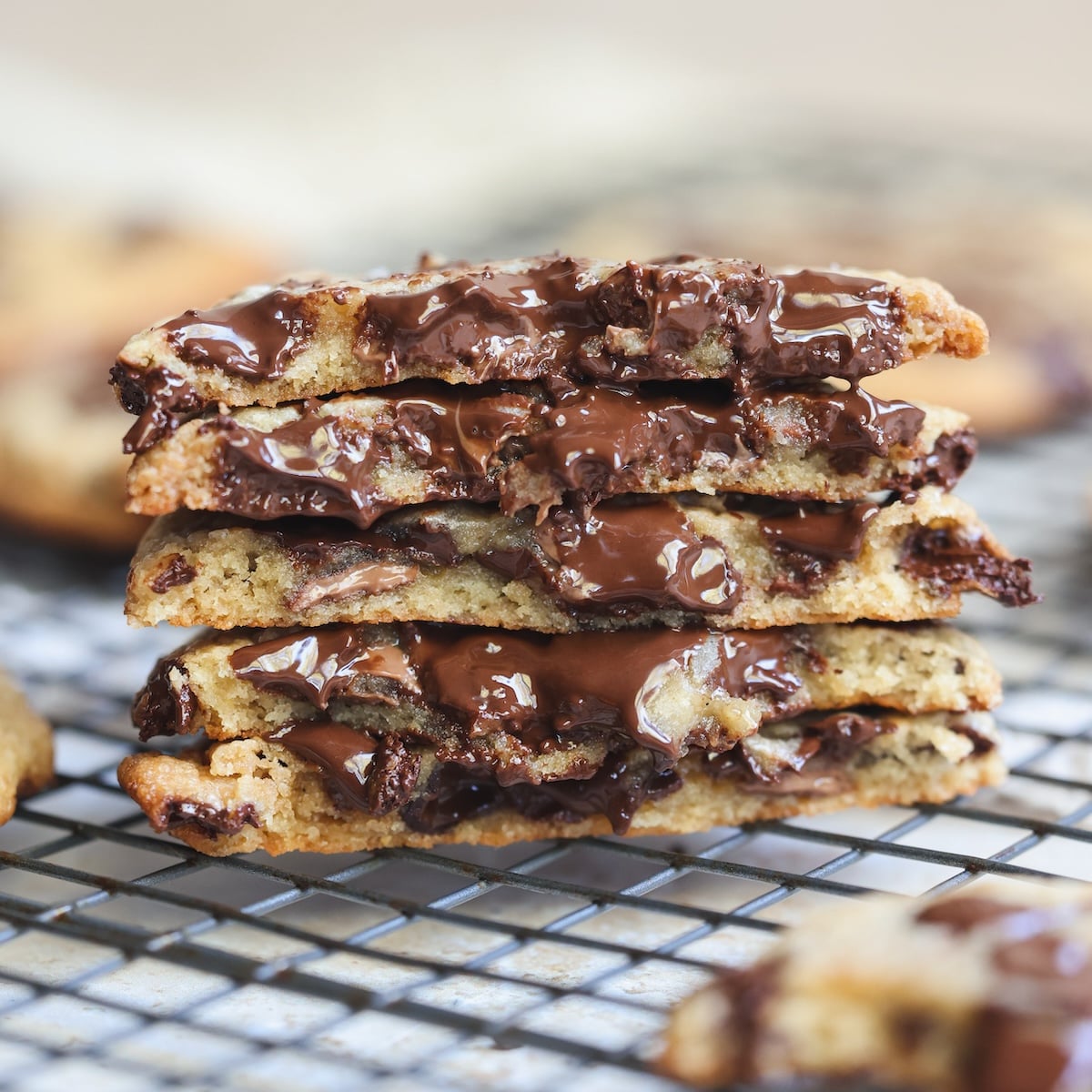 A stack of gooey chewy chocolate chip cookies, broken in half to show melted chocolate, sits on a wire cooling rack.