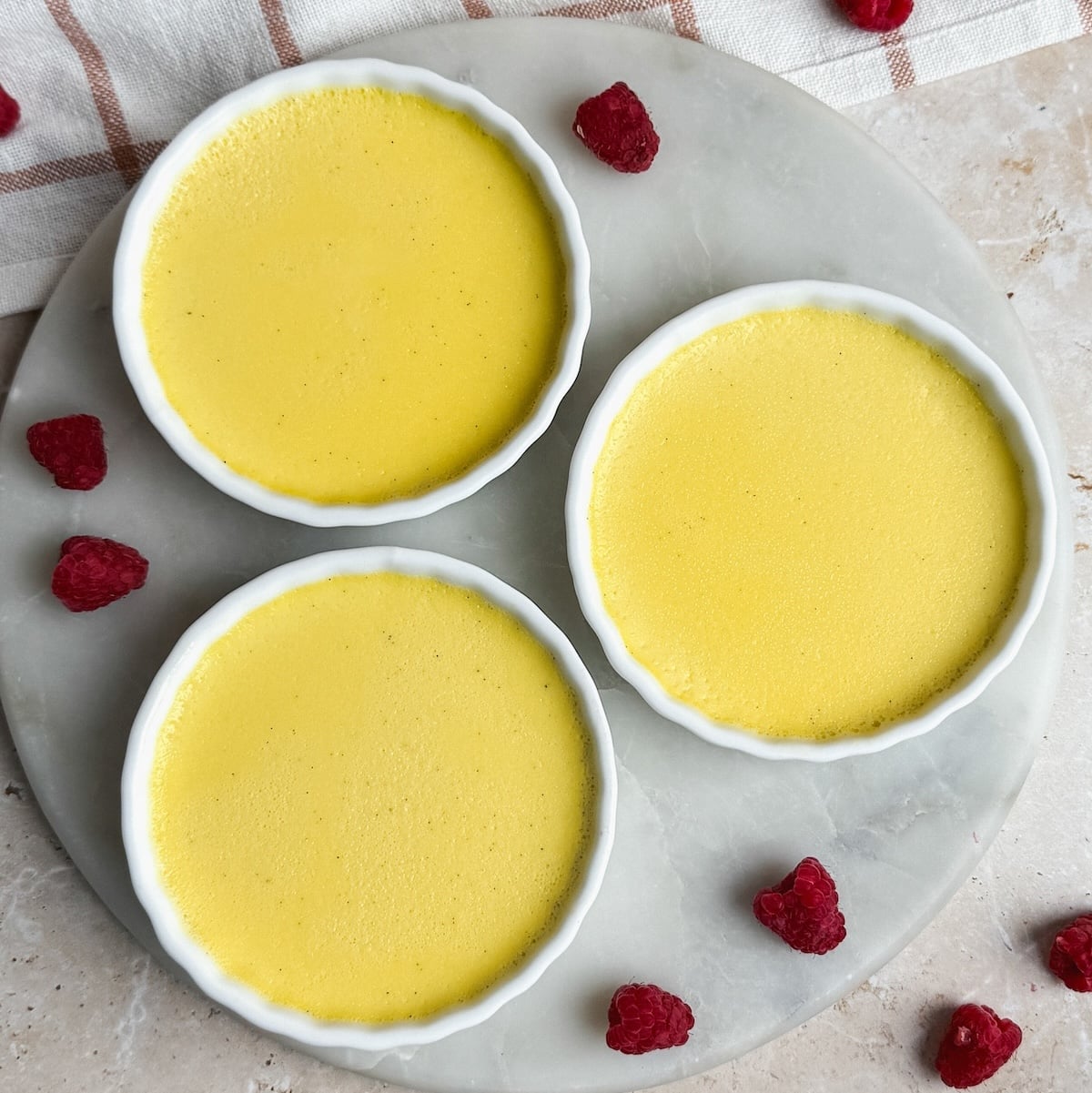Three small white ramekins filled with creamy yellow baked custard are arranged on a round marble surface, surrounded by scattered fresh raspberries. A striped towel is partially visible in the background.