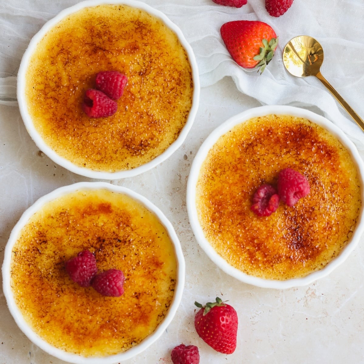 Three bowls of crème brûlée with caramelized golden tops, each garnished with raspberries. Fresh strawberries and a gold spoon are nearby on a light textured surface with a white cloth.