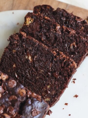 Three slices of moist and fudgy chocolate banana bread with chocolate chips, arranged on a white plate, with crumbs scattered around.