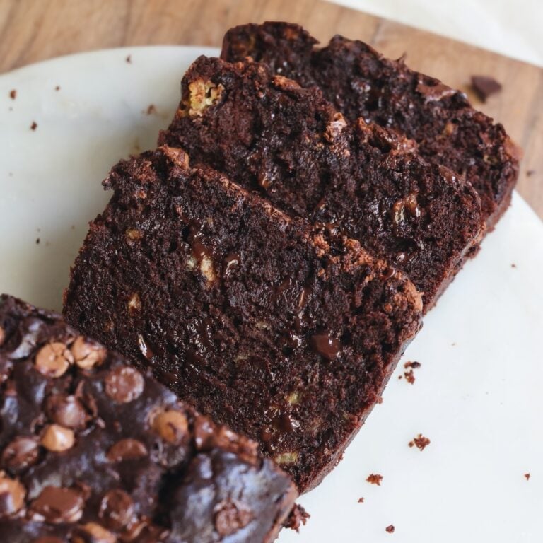 Three slices of moist and fudgy chocolate banana bread with chocolate chips, arranged on a white plate, with crumbs scattered around.