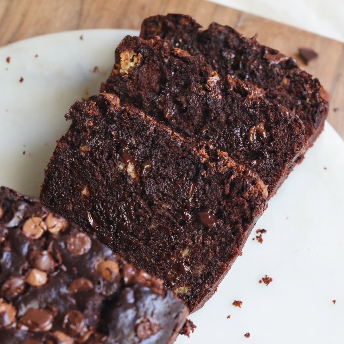 Three slices of moist and fudgy chocolate banana bread with chocolate chips, arranged on a white plate, with crumbs scattered around.