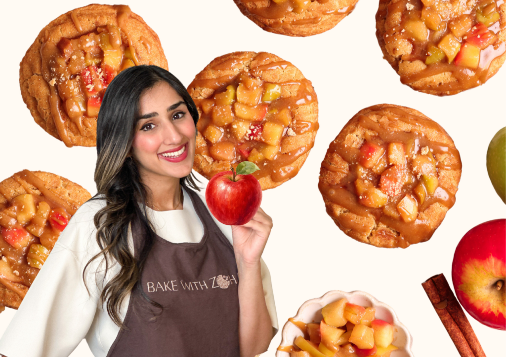 A smiling woman in a Bake With Zoha apron holds an apple, surrounded by several apple-topped cookies, apple slices, cinnamon sticks, and a bowl of apple filling on a light background—a delightful scene that captures the joy of baking with Zoha.