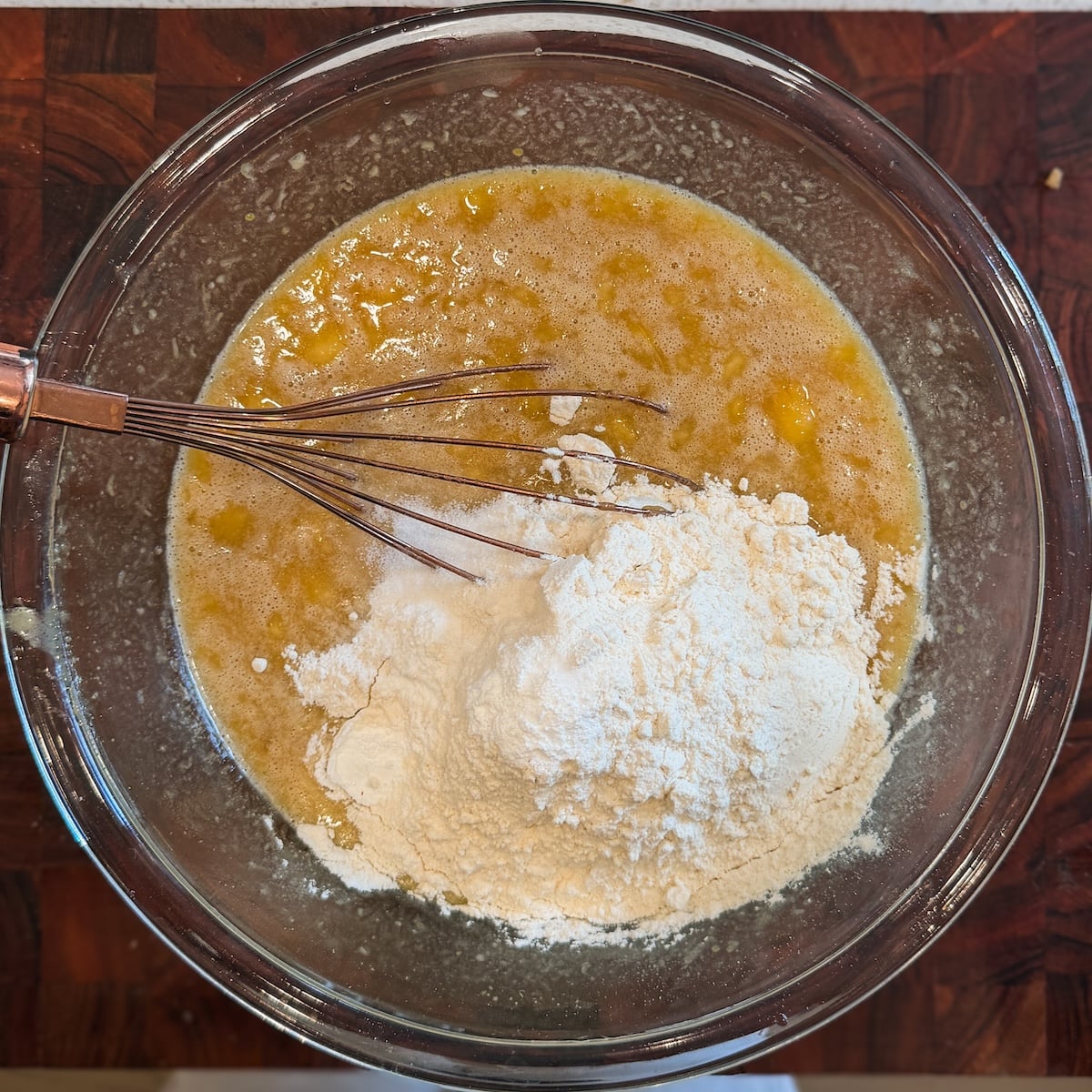 A glass bowl with partially mixed banana bread batter, including mashed bananas, flour, and wet ingredients. A metal whisk rests in the mixture atop a wooden countertop.