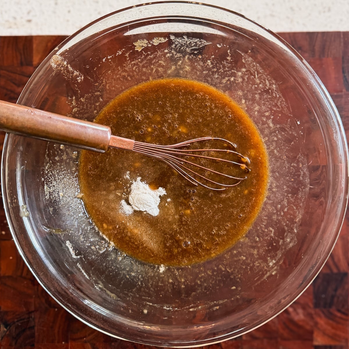 A glass bowl with brown liquid and a wire whisk rests on a wooden surface. Baking soda and salt added on top
