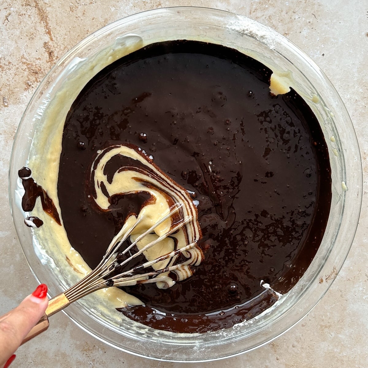 A hand holding a whisk stirs chocolate and vanilla batters together in a glass bowl, creating a marbled effect on a light countertop.