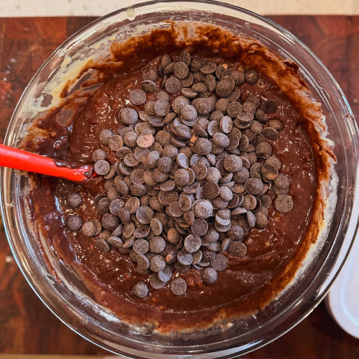 A glass bowl filled with chocolate banana bread batter and a pile of chocolate chips, being mixed with a red spatula, sits on a wooden surface.